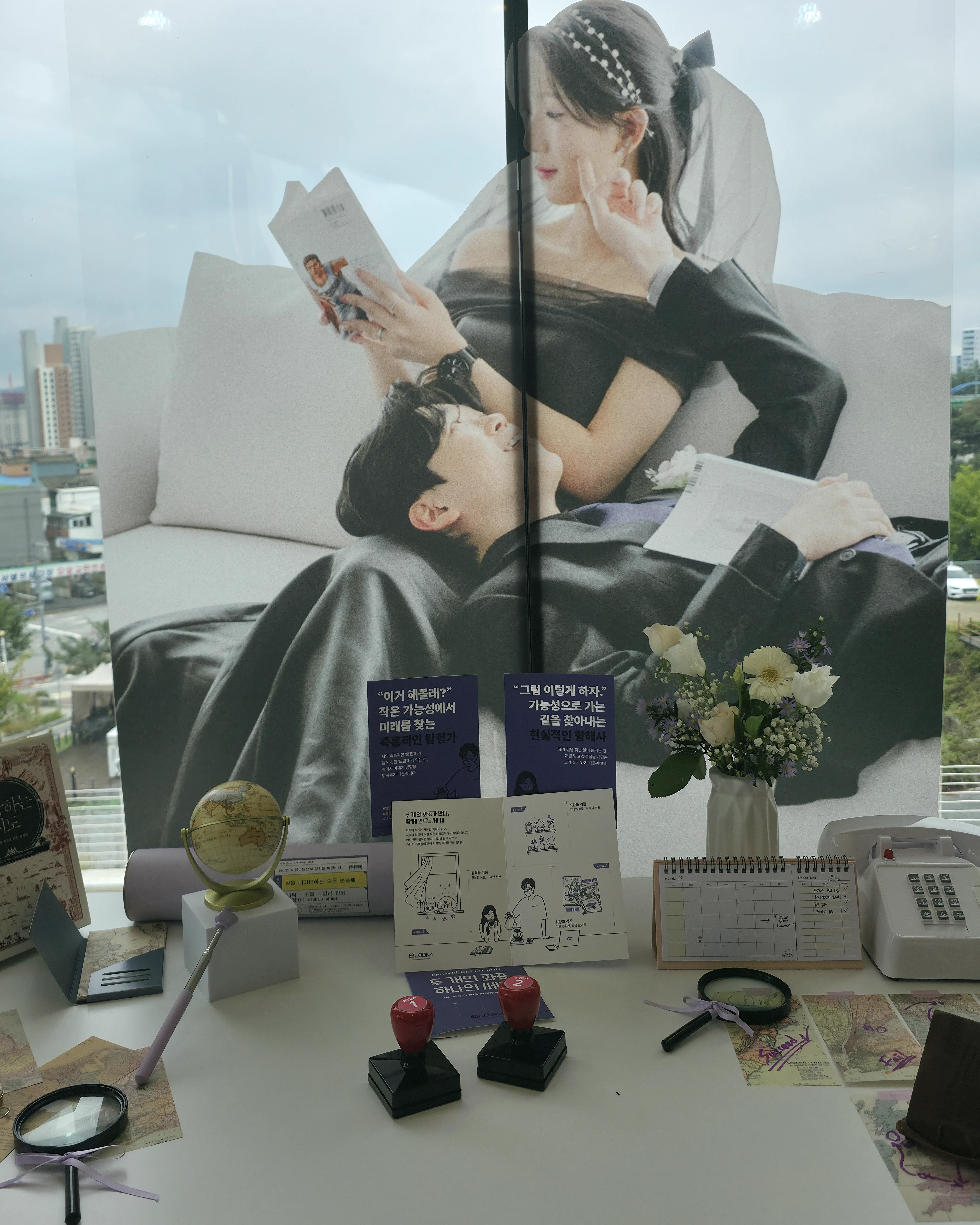 식장 아치창에 붙인 대형 사진 그래픽. 소파 위 두 사람 아래 창가 테이블에 보라 카드, 일러스트 리플렛, 빨간 도장 두 개, 돋보기, 유선전화