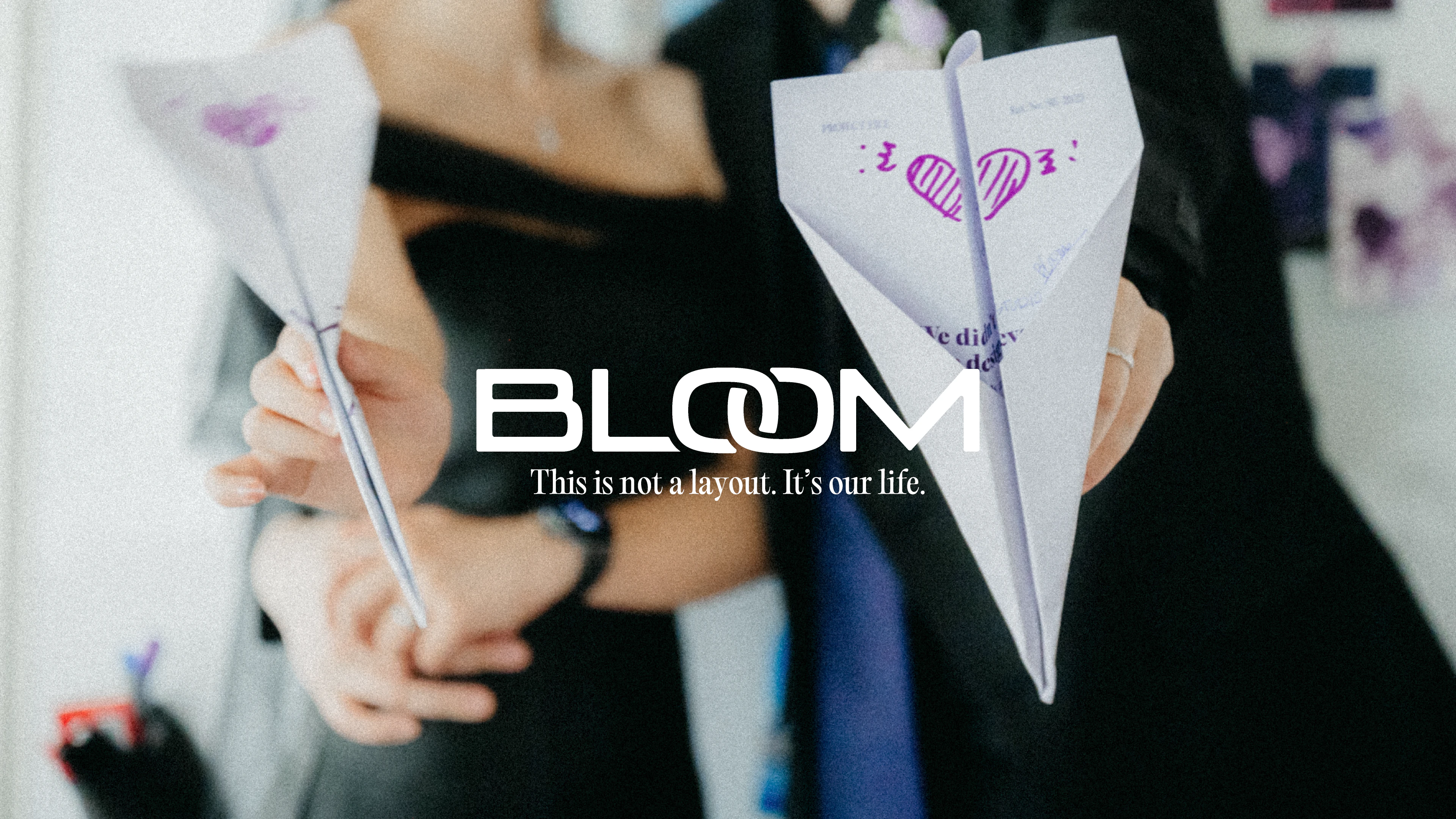 보라 하트 그래픽이 찍힌 종이비행기 두 개를 든 손. 사진 위에 BLOOM 워드마크와 "This is not a layout. It's our life." 문구.
