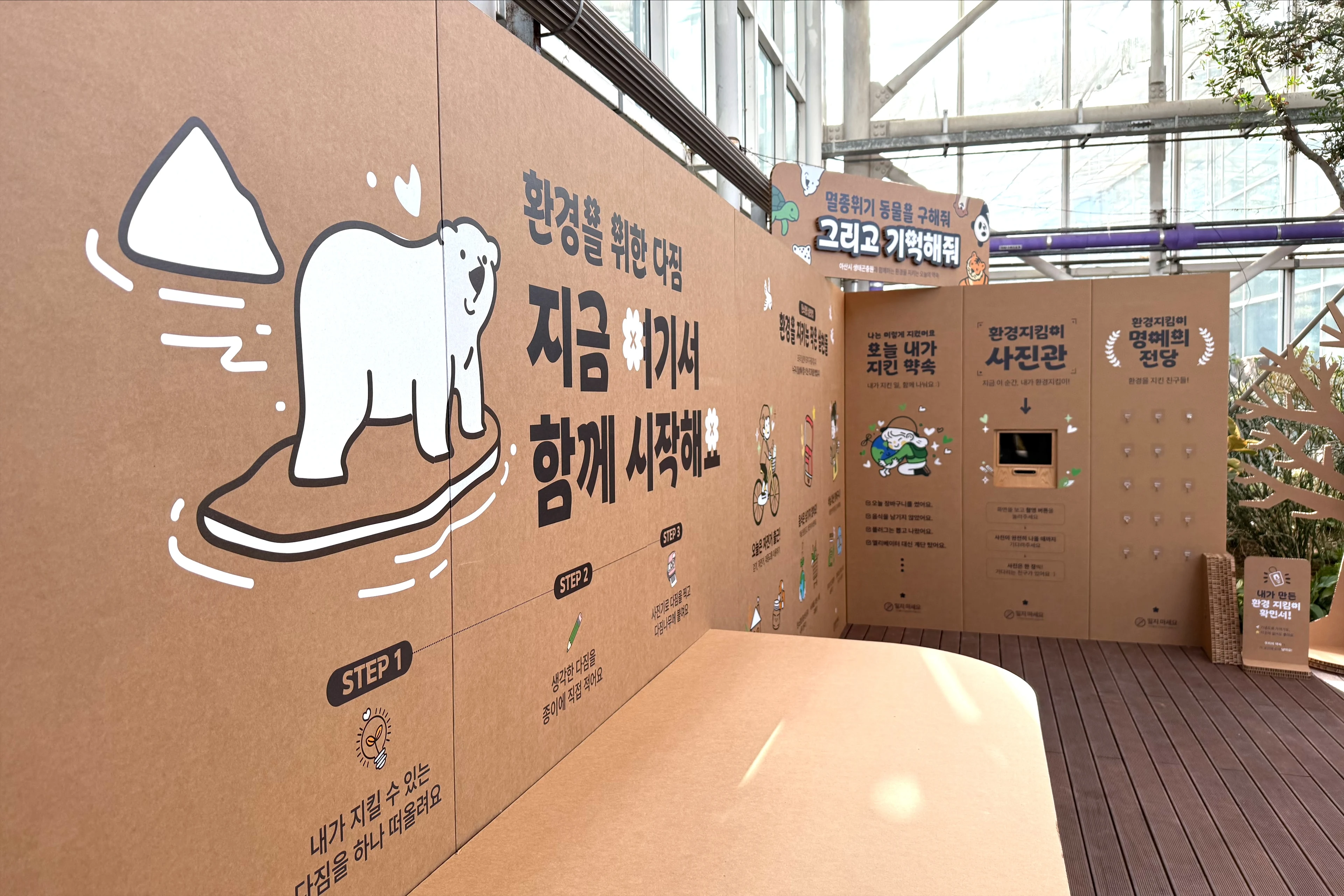 북극곰 일러스트와 '환경을 위한 다짐 — 지금 여기서 함께 시작해요' 문구가 있는 골판지 벽을 사선에서 본 모습