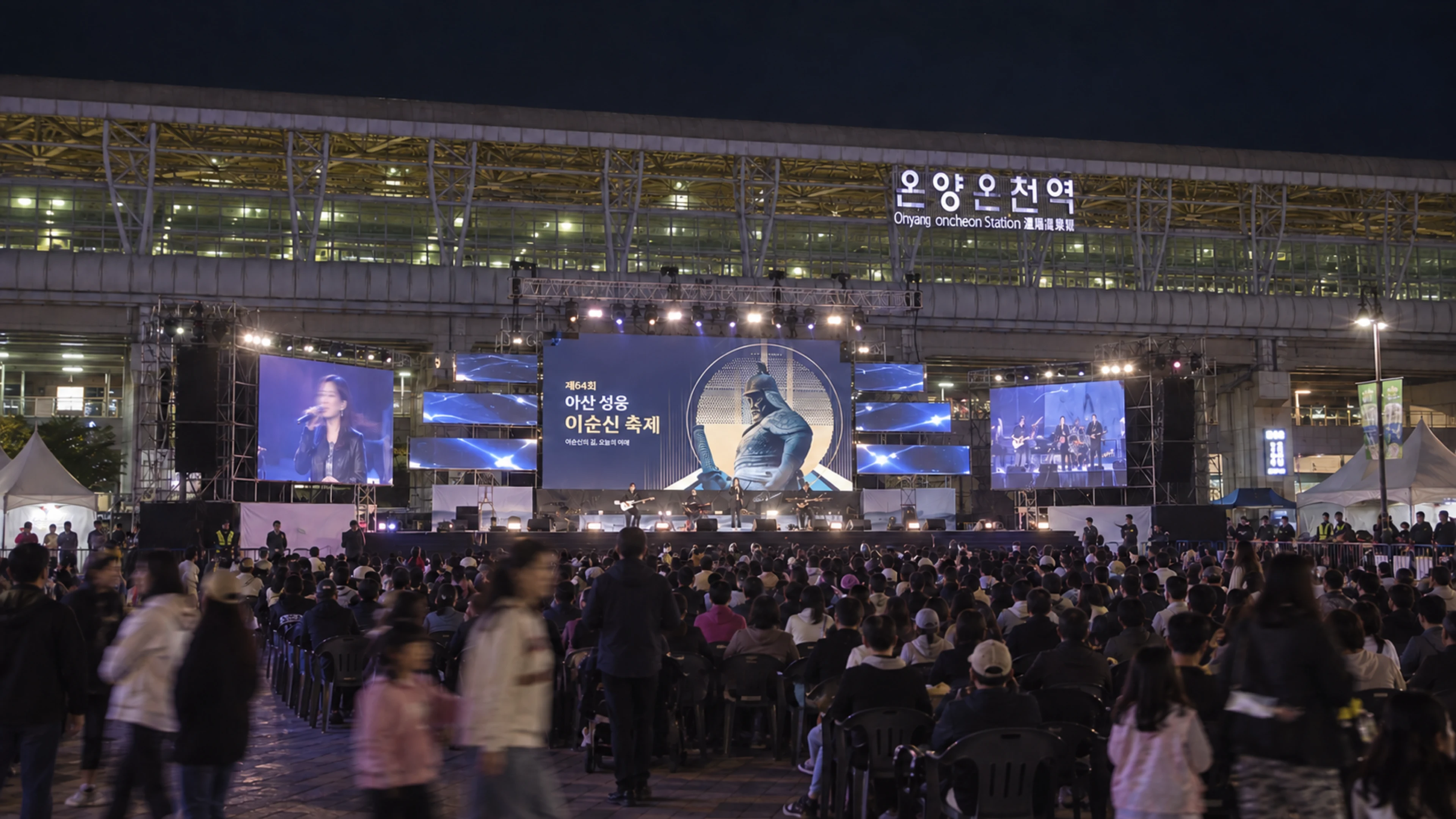 밤의 온양온천역 앞 축제 무대, 가운데 화면의 축제 그래픽과 객석의 관객 (AI로 구성한 이미지)
