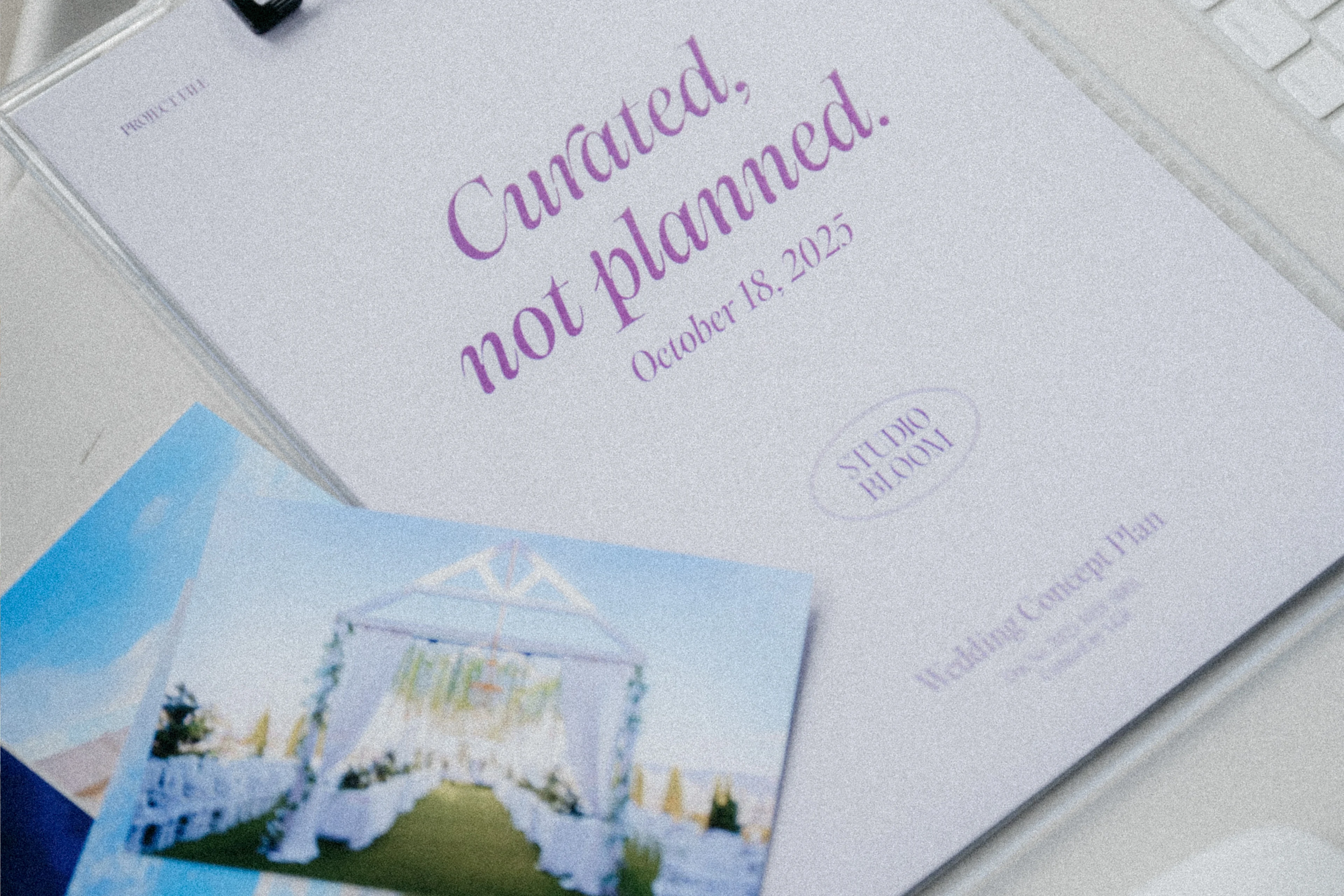 흰 책상 위 클립보드에 끼운 연보라 표지 기획서. "Curated, not planned. October 18, 2025"와 STUDIO BLOOM 타원 로고, 옆에 키보드·마우스와 식장 사진 카드