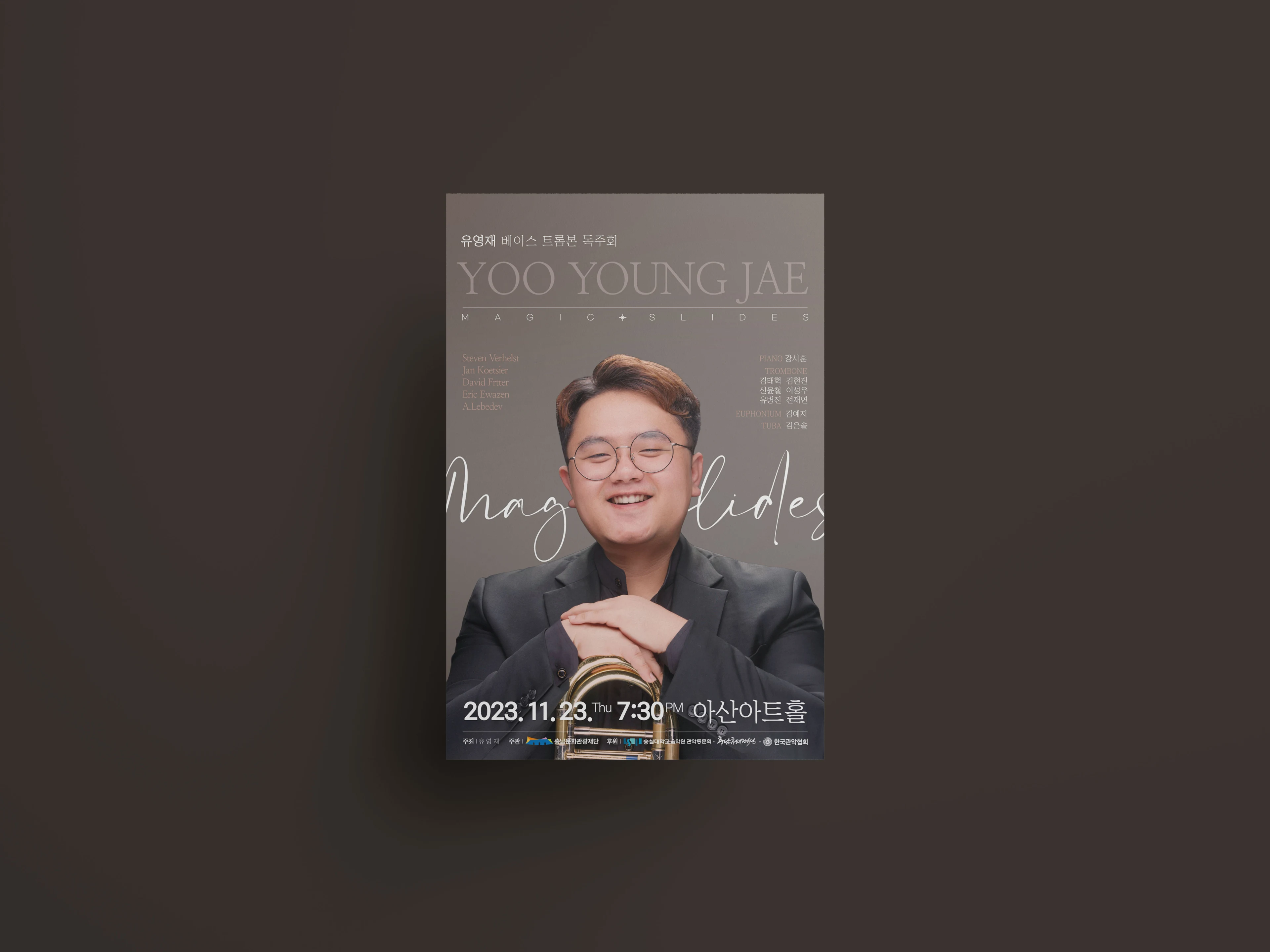 어두운 회갈색 벽에 붙은 〈MAGIC SLIDES〉 포스터. 트롬본에 손을 얹고 웃는 연주자와 필기체 공연명, 2023.11.23 아산아트홀 공연 정보.